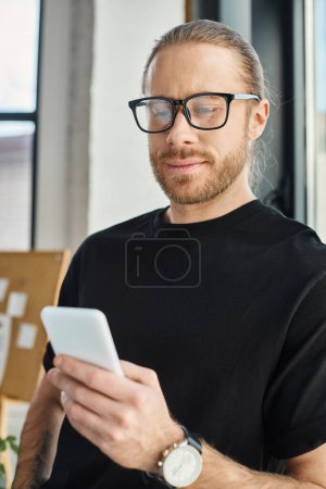 stylish businessman in black t-shirt and eyeglasses browsing internet on smartphone in office