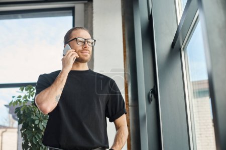 hombre de negocios seguro de sí mismo en camiseta negra y anteojos hablando en el teléfono inteligente y mirando hacia otro lado