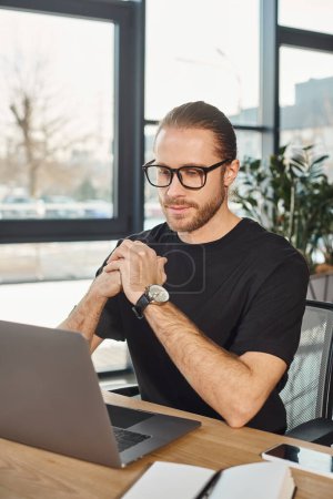 thoughtful businessman in black t-shirt and eyeglasses looking at laptop at workplace in office