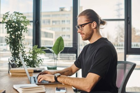 hombre de negocios sonriente en camiseta negra y anteojos escribiendo en el ordenador portátil en el lugar de trabajo en la oficina moderna