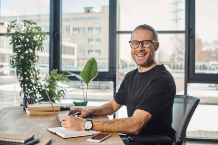 joyful businessman in black t-shirt and eyeglasses looking at camera near laptop on work desk