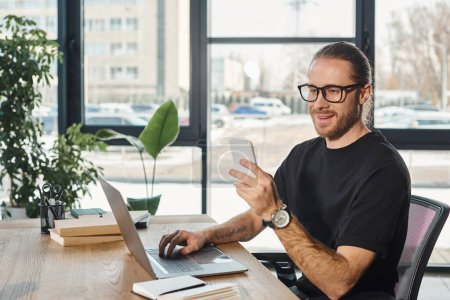 happy businessman in eyeglasses looking at smartphone near laptop on work desk in modern office