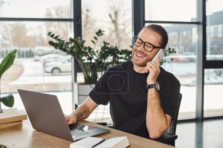 fröhlicher Geschäftsmann mit Brille, der am Arbeitsplatz im modernen Büro auf dem Smartphone neben dem Laptop spricht