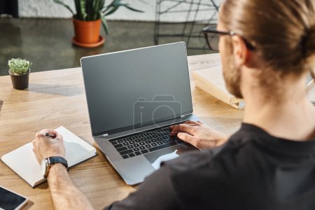businessman in black t-shirt typing on laptop with blank screen and writing in notebook, back view