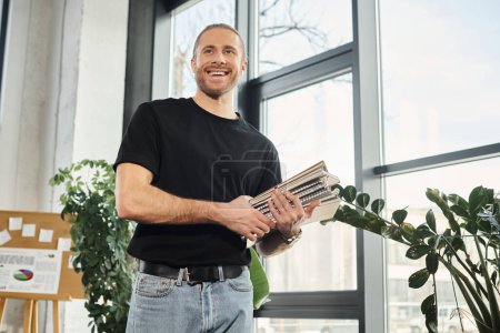 alegre hombre de negocios en camiseta negra sosteniendo pila de cuadernos y mirando hacia otro lado en la oficina moderna