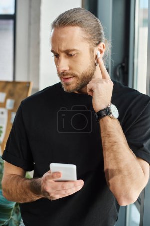 thoughtful businessman with smartphone adjusting earphone while listening podcast in office