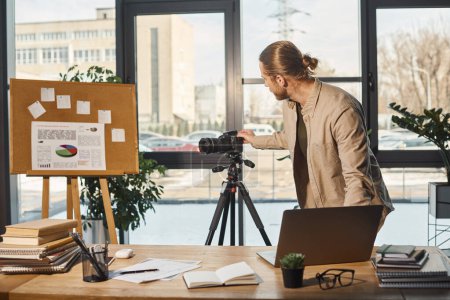 Photo for Corporate manager adjusting digital camera on tripod near flip chart with graphs and office desk - Royalty Free Image