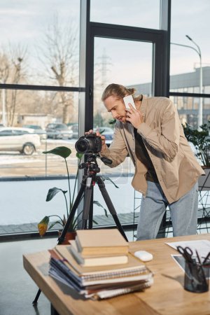 Foto de Hombre de negocios hablando en el teléfono inteligente y ajustando la cámara digital junto al escritorio en la oficina moderna - Imagen libre de derechos