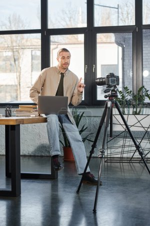 businessman sitting on work desk with laptop and talking in front of digital camera, video blog