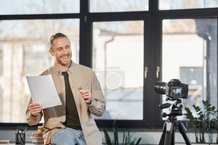 smiling entrepreneur holding bitcoin and document in front of digital camera in modern office