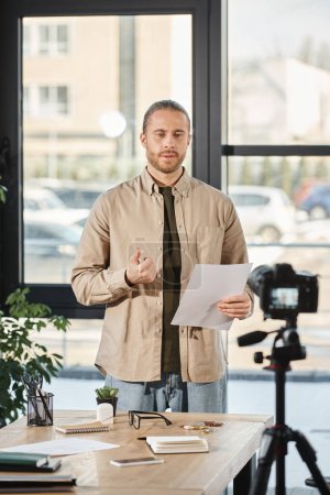Foto de Hombre de negocios serio sosteniendo documento y hablando delante de la cámara digital en la oficina moderna - Imagen libre de derechos