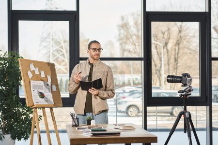 Geschäftsmann mit Brille hält Notizbuch in der Hand und spricht bei Videoblog im Büro an Digitalkamera