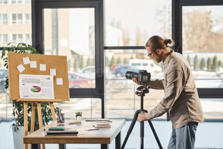 Unternehmer in Freizeitkleidung stellt Digitalkamera neben Schreibtisch und Korkboard mit Diagrammen im Büro ein
