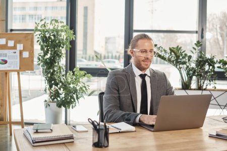 positive businessman in grey suit and eyeglasses working on laptop at workplace in modern office