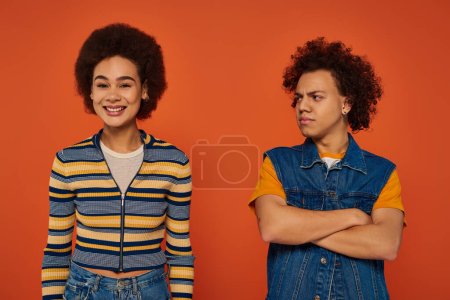 young african american man looking dissatisfied while sister smiling cheerfully on orange backdrop