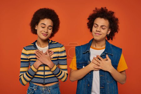 Zufriedene afrikanisch-amerikanische Geschwister posieren mit den Händen auf der Brust mit geschlossenen Augen vor orangefarbenem Hintergrund