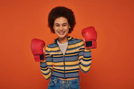 beautiful jolly african american woman posing actively with boxing gloves on orange backdrop