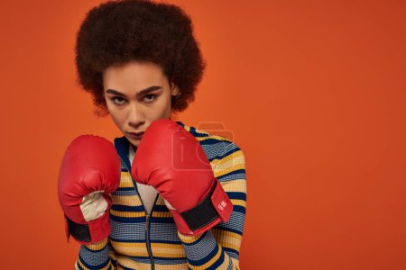 beautiful focused african american woman posing actively with boxing gloves on orange backdrop