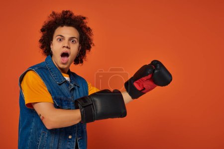 modelo masculino afroamericano deportivo sorprendido posando animado en guantes de boxeo sobre fondo naranja