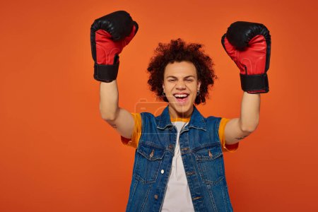 guapo alegre afroamericano hombre posando activamente en guantes de boxeo sobre fondo naranja