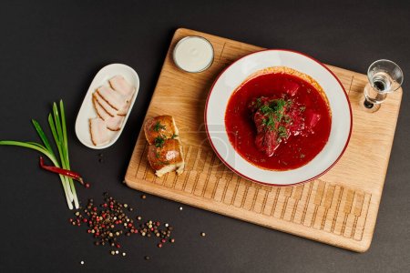 Ukrainische Rote-Bete-Suppe in der Nähe von Knoblauchbrötchen, Schweineschmalz und grünen Zwiebeln auf Schneidebrett auf Schwarz
