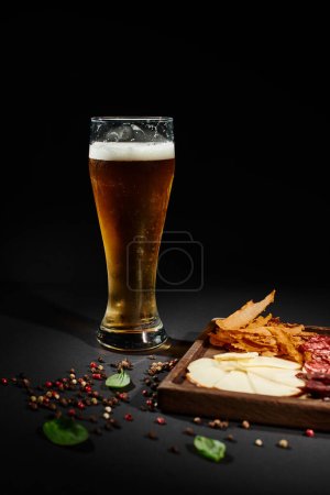 glass of craft beer near charcuterie board with gourmet cheese, dried beef and salami on black
