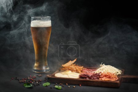 glass of beer near charcuterie board with gourmet cheese, dried beef and salami on black backdrop