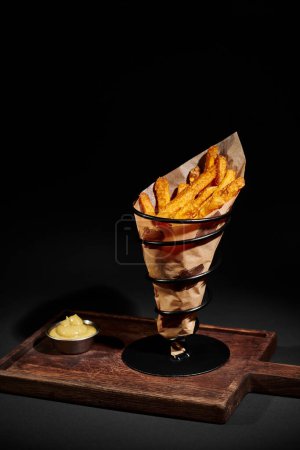 Photo for Delicious crispy French fries inside of paper cone near dipping sauce on wooden cutting board - Royalty Free Image