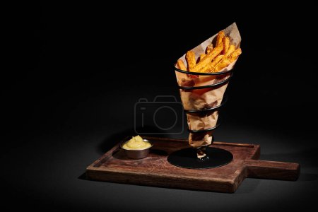 Téléchargez les photos : Délicieuses et croustillantes frites à l'intérieur du cône de papier près de sauce à tremper sur planche à découper en bois - en image libre de droit