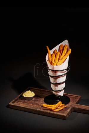 Téléchargez les photos : Savoureuses frites croustillantes à l'intérieur du cône de papier près de sauce à tremper sur planche à découper en bois - en image libre de droit