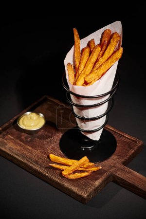 Photo for Cooked and crispy French fries inside of paper cone near dipping sauce on wooden cutting board - Royalty Free Image