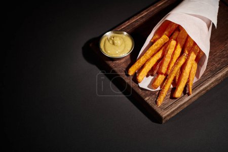 Photo for Delicious homemade French fries inside of paper cone near dipping sauce on wooden cutting board - Royalty Free Image