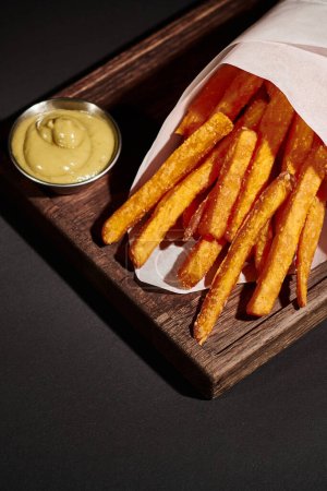 Photo for Vertical shot of crispy French fries inside of paper cone near dipping sauce on wooden cutting board - Royalty Free Image
