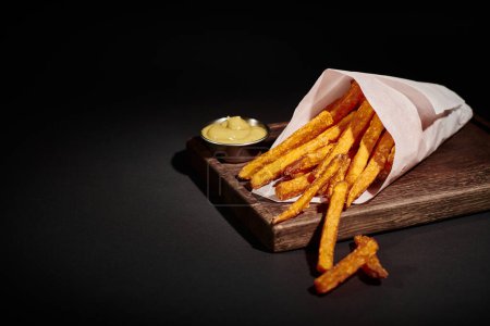 Photo for Quick meal, crispy French fries inside of paper cone near dipping sauce on wooden cutting board - Royalty Free Image