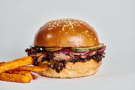tasty hamburger with beef, red onion, tomato and sesame bun near French fries on grey backdrop