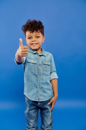 Téléchargez les photos : Frisé afro-américain préscolaire garçon en tenue de denim élégant montrant comme sur fond bleu - en image libre de droit