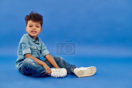 Téléchargez les photos : Garçon préscolaire afro-américain souriant en denim élégant assis sur fond bleu - en image libre de droit