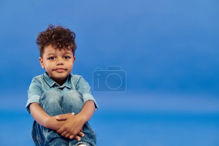 Téléchargez les photos : Garçon préscolaire afro-américain positif en denim élégant assis sur fond bleu - en image libre de droit