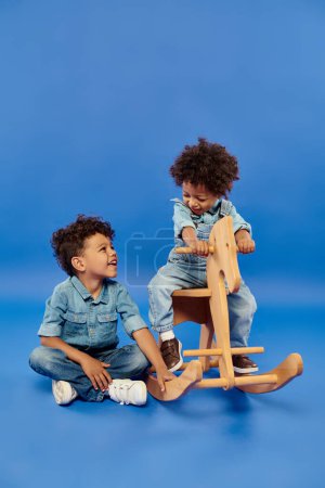 cute african american toddler boy in stylish denim clothes sitting on rocking horse near brother