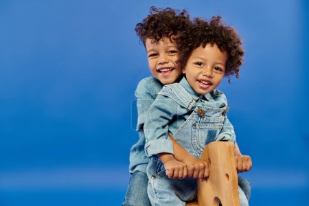 happy african american brothers in stylish denim clothes sitting on rocking horse on blue backdrop
