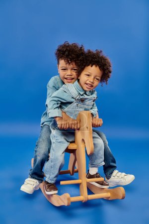happy african american boys in stylish denim clothes sitting on rocking horse on blue backdrop