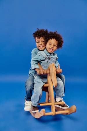 happy african american siblings in denim clothes sitting together on rocking horse on blue backdrop