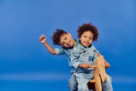 cute african american siblings in denim clothes sitting together on rocking horse on blue backdrop