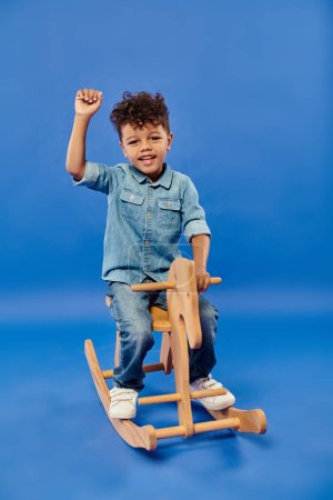 lindo africano americano preescolar chico en elegante denim ropa sentado en balanceo caballo en azul