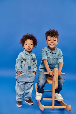 cute african american toddler boy in stylish denim clothes standing near brother on rocking horse