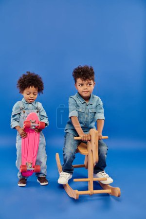 Photo for African american boy in stylish denim clothes holding penny board near brother on rocking horse - Royalty Free Image