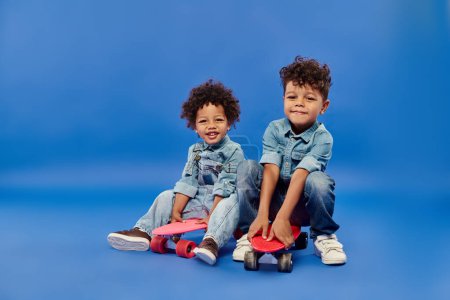 happy african american siblings in stylish denim clothes sitting on penny boards on blue backdrop