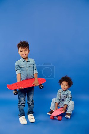 cute african american siblings in stylish denim clothes with penny boards on blue backdrop