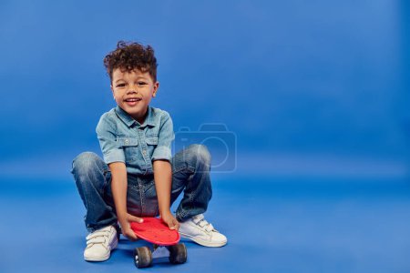 Téléchargez les photos : Mignon garçon préscolaire afro-américain en denim élégant assis sur un panneau de penny sur bleu - en image libre de droit