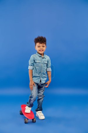Téléchargez les photos : Mignon garçon préscolaire afro-américain dans des vêtements en denim élégant debout sur le panneau penny sur bleu - en image libre de droit